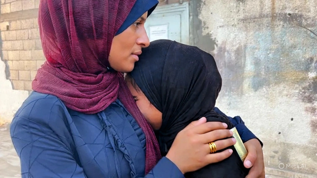 Two older women are shown close-up. The woman on the left has wrinkles and a distressed expression, wearing a pink and white head covering. The woman on the right wears a black niqab, revealing only her eyes, which appear weary or sad. In the blurred background, another woman in a white hijab can be seen. The setting appears to be outdoors, possibly near temporary structures.