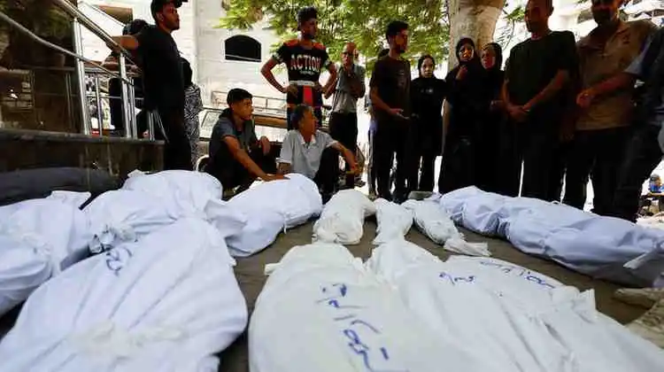 The image depicts a somber scene where a group of people, likely family members or mourners, gather around a truck loaded with white shrouds, each bearing a blue logo. The atmosphere is one of grief and solemnity, as the individuals stand in contemplative silence, their faces reflecting a mix of sadness and reverence. The presence of the shrouds suggests that the occasion is a funeral or memorial service, and the logo on the shrouds may indicate that the deceased were part of a particular group or organization. The overall mood is subdued, with the natural surroundings, including trees and buildings in the background, providing a stark contrast to the somber mood of the gathering.